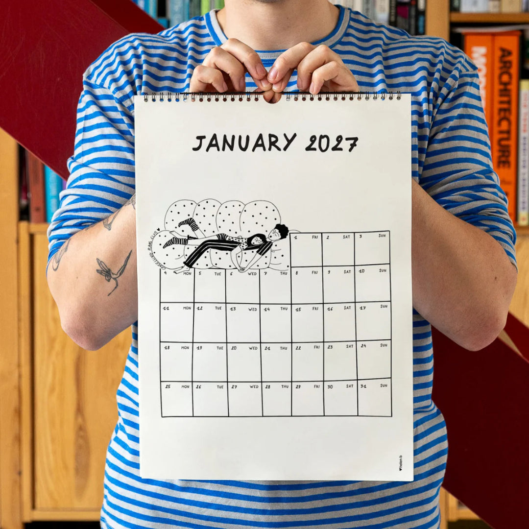 Chico con camiseta a rayas azules sosteniendo el calendario 2027 ilustrado por Helen B abierto por el mes de enero, con ilustración de una pareja tumbada y dormida sobre un sofá mullido de puntos.