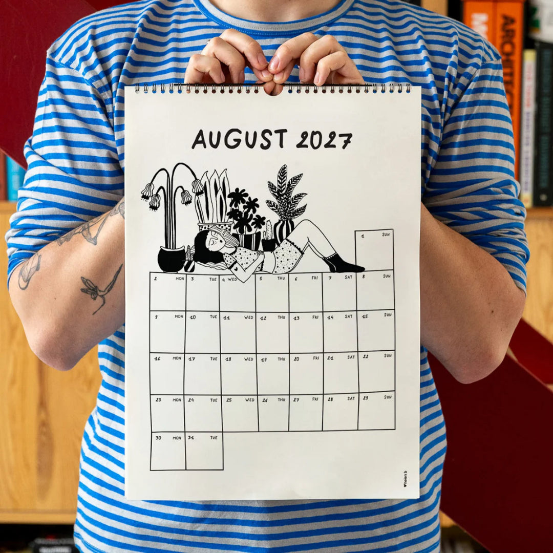 Chico con camiseta a rayas azules sosteniendo el calendario 2027 ilustrado por Helen B abierto por el mes de enero, con ilustración de una chica dormida junto a varias macetas con plantas