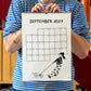 Chico con camiseta a rayas azules sosteniendo el calendario 2027 ilustrado por Helen B abierto por el mes de septiembre, con ilustración de una chica tirando de la correa de un perro que no quiere moverse