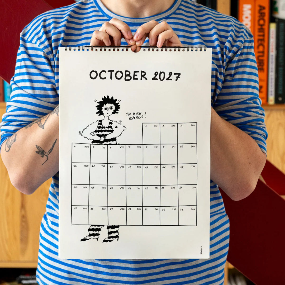 Chico con camiseta a rayas azules sosteniendo el calendario 2027 ilustrado por Helen B abierto por el mes de octubre, con ilustración de una chica temblando porque ha tomado demasiado café