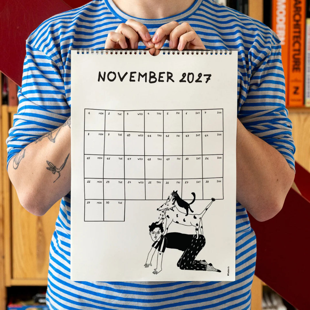 Chico con camiseta a rayas azules sosteniendo el calendario 2027 ilustrado por Helen B abierto por el mes de noviembre, con ilustración de un chico a cuatro patas con un perro encima y un gato encima del perro