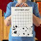 Chico con camiseta a rayas azules sosteniendo el calendario 2027 ilustrado por Helen B abierto por el mes de diciembre, con ilustración de Papá Noël corriendo detrás de un reno que lleva sus calzoncillos en los cuernos