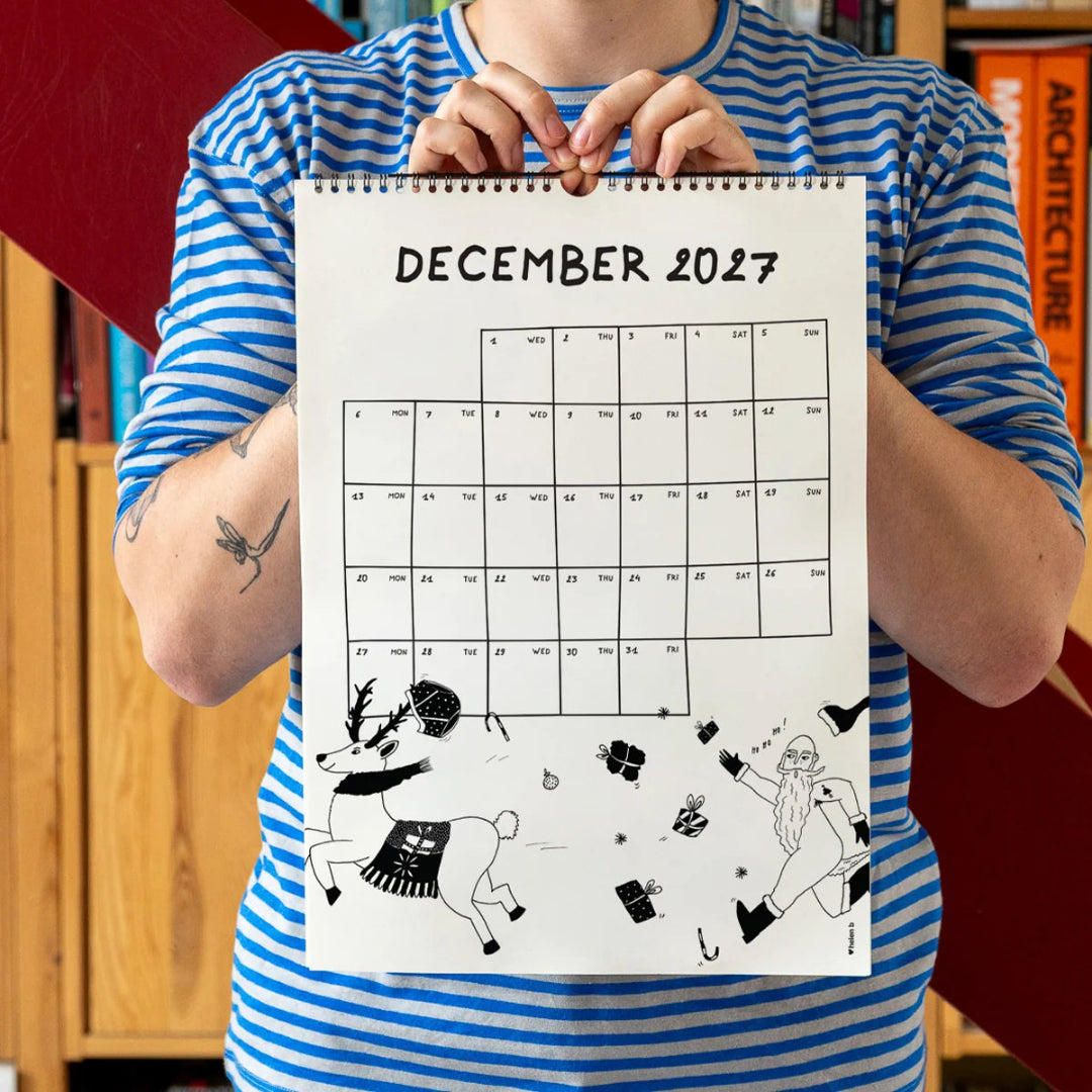 Chico con camiseta a rayas azules sosteniendo el calendario 2027 ilustrado por Helen B abierto por el mes de diciembre, con ilustración de Papá Noël corriendo detrás de un reno que lleva sus calzoncillos en los cuernos