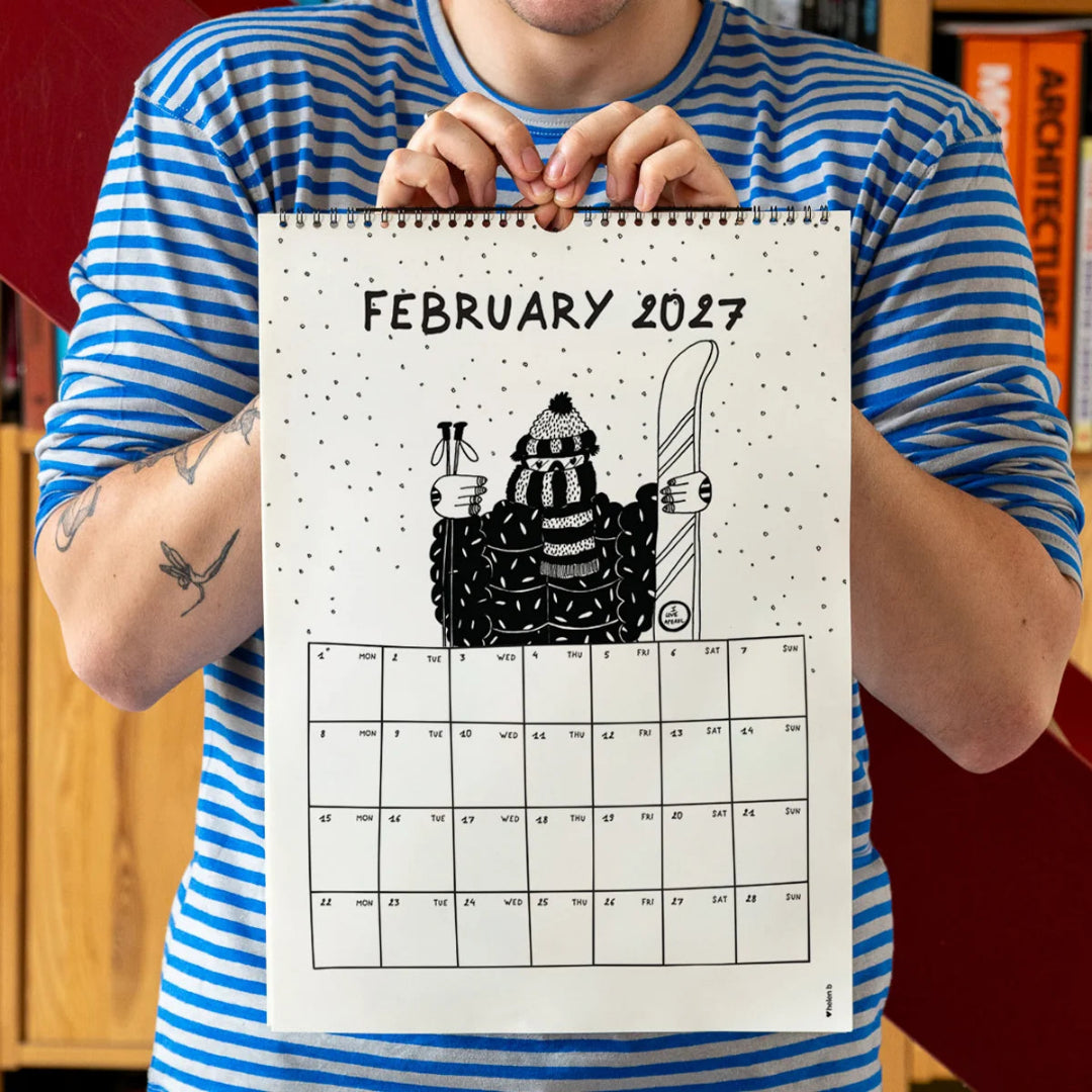 Chico con camiseta a rayas azules sosteniendo el calendario 2027 ilustrado por Helen B abierto por el mes de febrero, con ilustración de una esquiadora