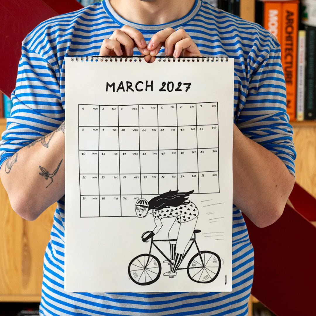 Chico con camiseta a rayas azules sosteniendo el calendario 2027 ilustrado por Helen B abierto por el mes de marzo, con ilustración de una ciclista