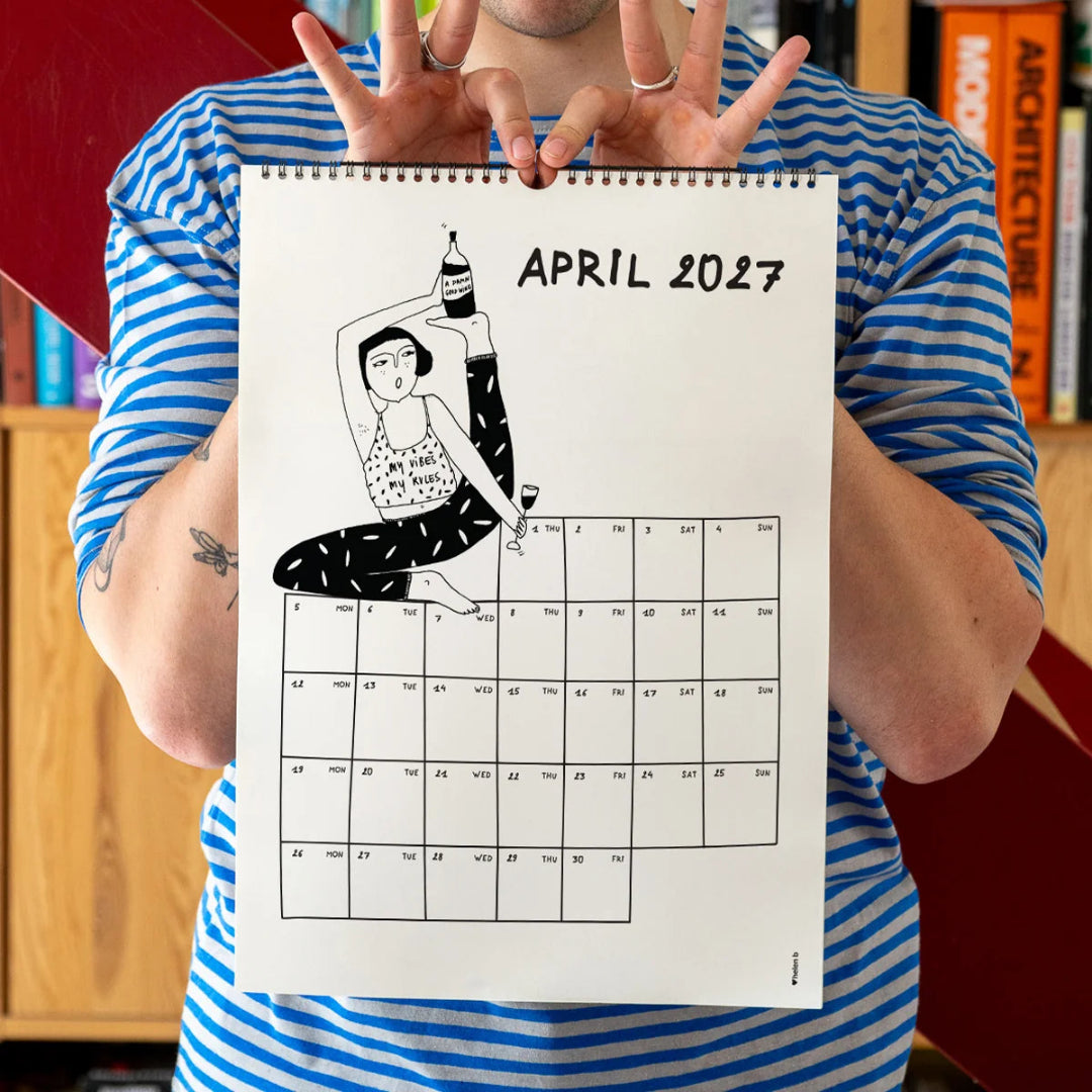 Chico con camiseta a rayas azules sosteniendo el calendario 2027 ilustrado por Helen B abierto por el mes de abril, con ilustración de una chica practicando yoga con una botella de vino