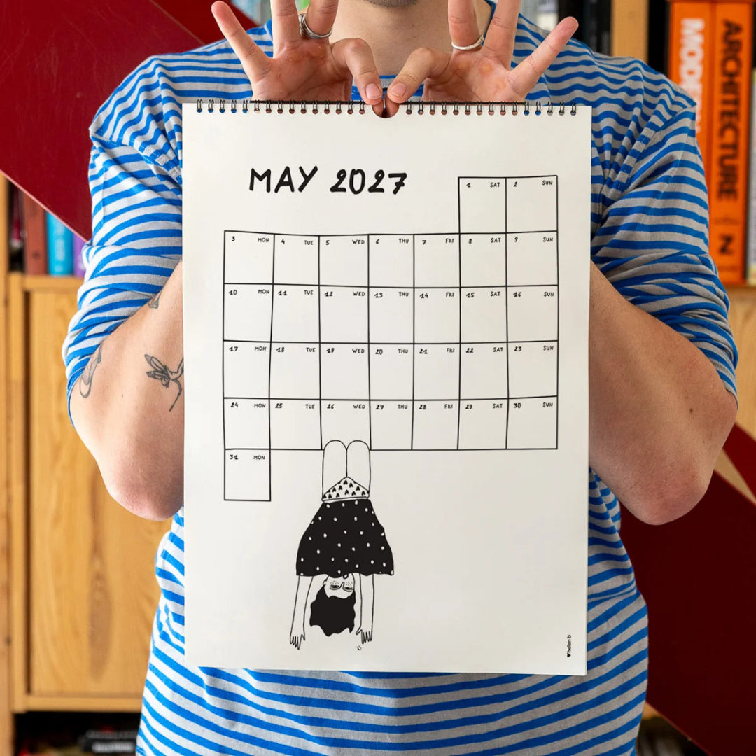 Chico con camiseta a rayas azules sosteniendo el calendario 2027 ilustrado por Helen B abierto por el mes de mayo, con ilustración de una trapecista con la falda del revés y braguitas de corazones
