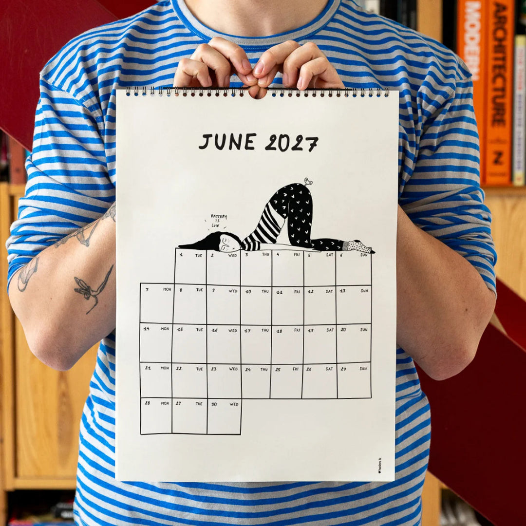 Chico con camiseta a rayas azules sosteniendo el calendario 2027 ilustrado por Helen B abierto por el mes de junio, con ilustración de una chica dormida con el culo en pompas