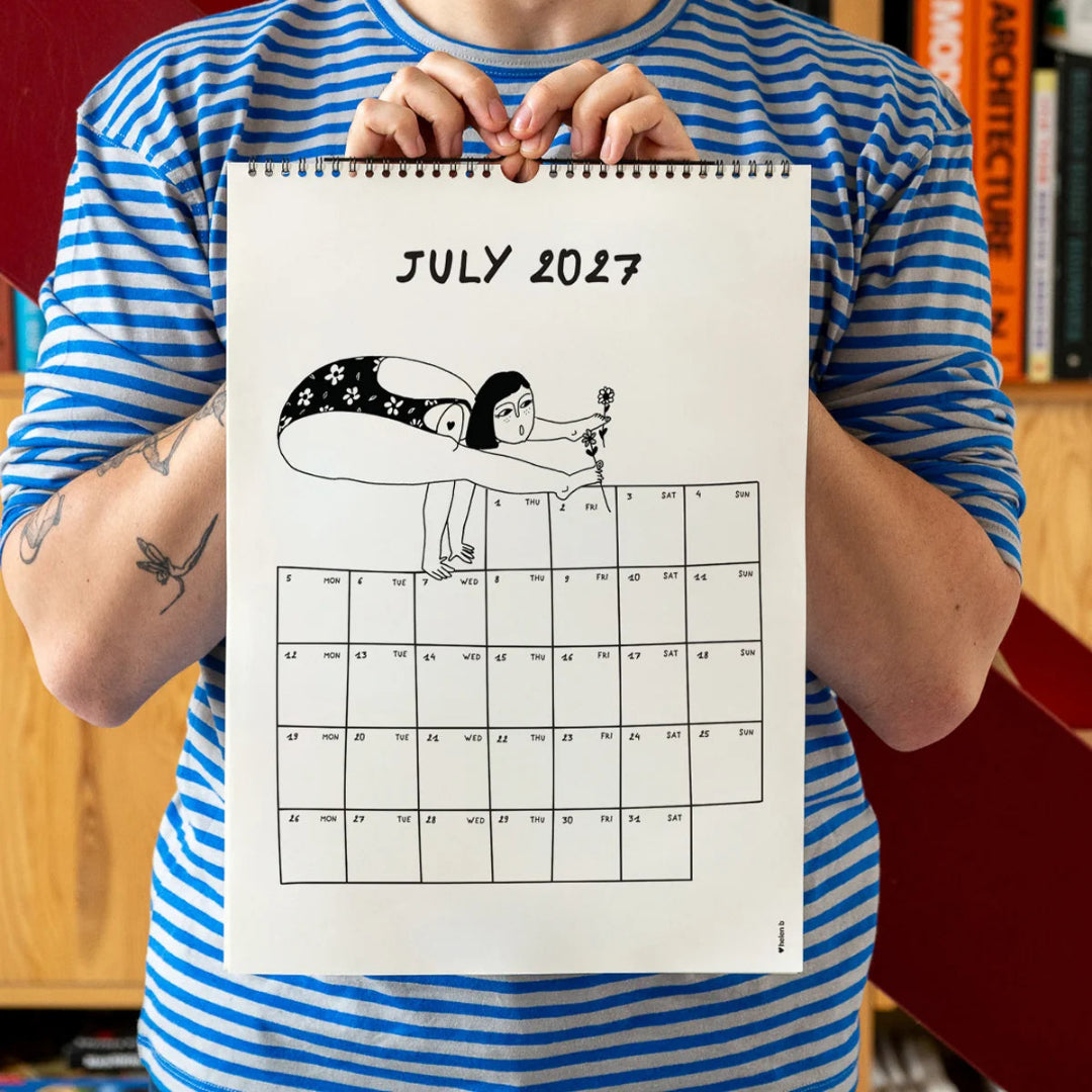 Chico con camiseta a rayas azules sosteniendo el calendario 2027 ilustrado por Helen B abierto por el mes de julio, con ilustración de una chica en bañador en postura acrobática sosteniendo unas flores con los pies