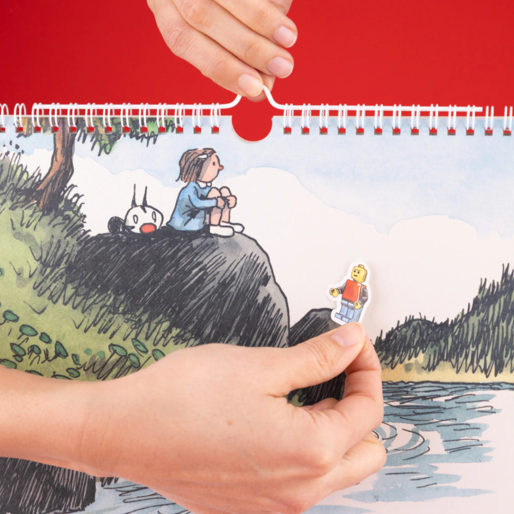 Detalle del Calendario de pared 2027 de Macanudo con una mano pegando una pegatina sobre una de las ilustraciones