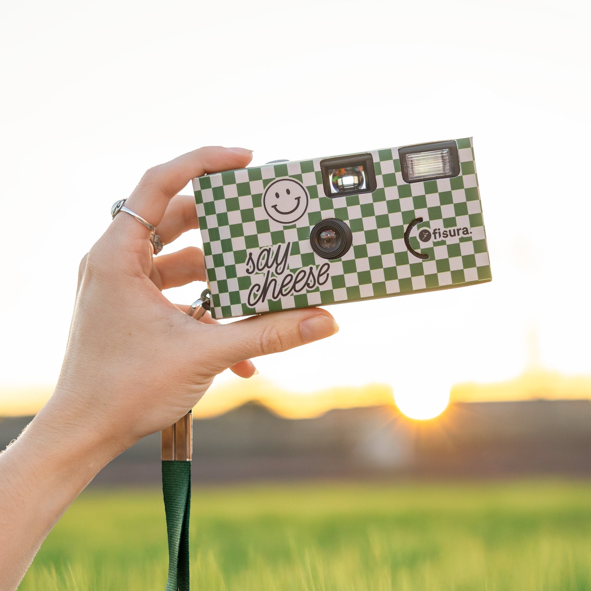 Mujer sosteniendo una cámara desechable con aires retro a cuadros verdes y beiges con correa para colgar en un campo verde con el atardecer de fondo
