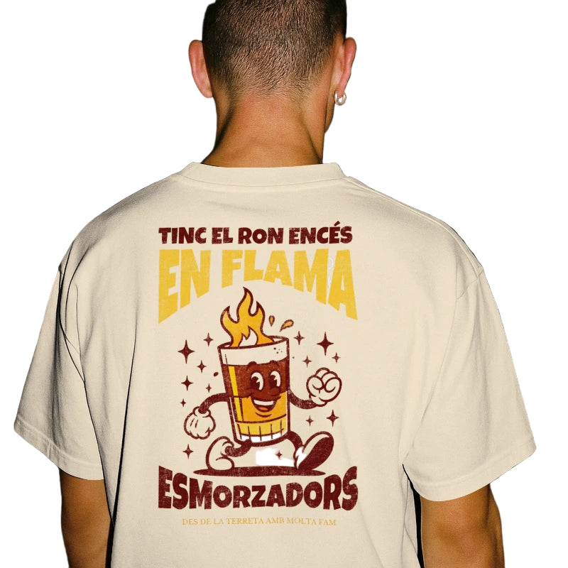 Chico de espaldas con la camiseta de Esmorzadors Cremaet, con un vaso de café con ron andando y la frase "tinc el ron encés en flama"