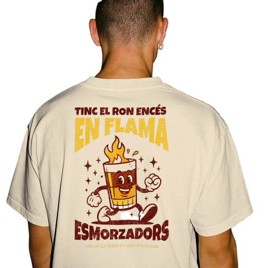 Chico de espaldas con la camiseta de Esmorzadors Cremaet, con un vaso de café con ron andando y la frase "tinc el ron encés en flama"