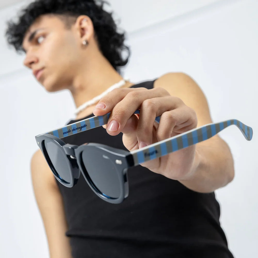 modelo sujetando con la mano las gafas de sol de Fisura redondas en color negro con las patillas a rayas azules y marrón, del modelo "Modo Chiringuito"