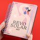 Portada de la Agenda Astrológica 2027 Cisne Revo Solar de Dominio Salvaje con tapa rosa, ilustración de cisne estilizado, espiral azul y elástico de cierre, sostenida por dos manos sobre fondo rojo
