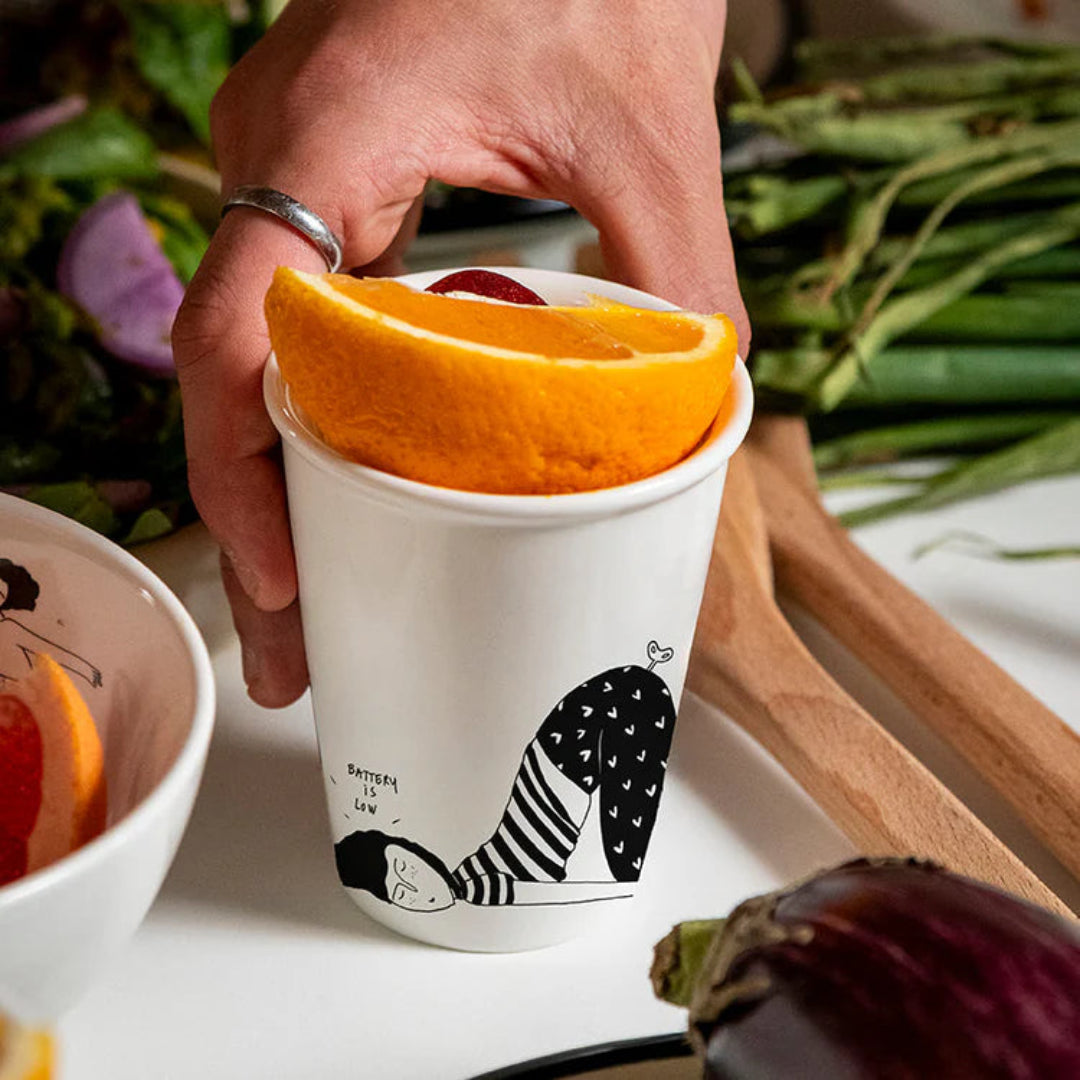 Taza blanca con un dibujo negro de una mujer durmiendo con el letrero "Battery is low", con una naranja dentro y una mano sujetando la taza.