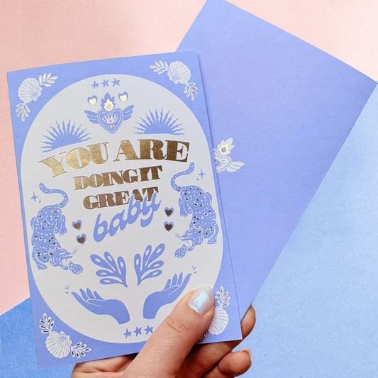 mano sujetando la tarjeta de la ilustradora Laura Ortiz con el texto "you are doing great baby" en color azul y con dibujos de dos manos, dos leopardos, un corazón con llamas y conchas de mar