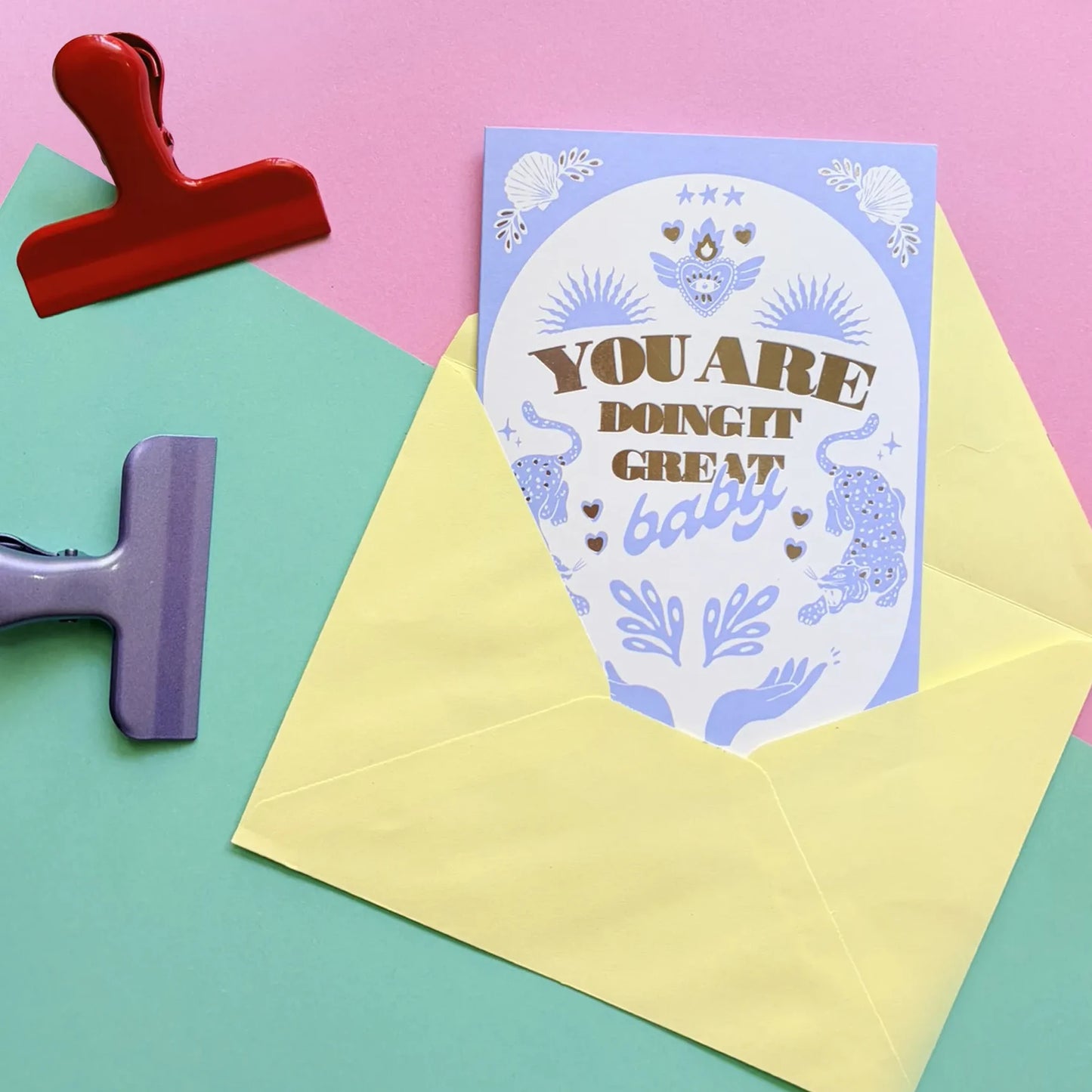 tarjeta de la ilustradora Laura Ortiz con el texto "you are doing great baby" en color azul y con dibujos de dos manos, dos leopardos, un corazón con llamas y conchas de mar, saliendo de un sobre de color amarillo y con dos pinzas a los lados