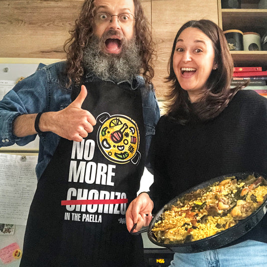 dos personas contentas en una cocina preparando la comida, una de ellas con el delantal en color negro de jajajers con el texto en blanco No more chorizo in the paella, y con la palabra chorizo tachada en rojo y un dibujo de una paella al lado