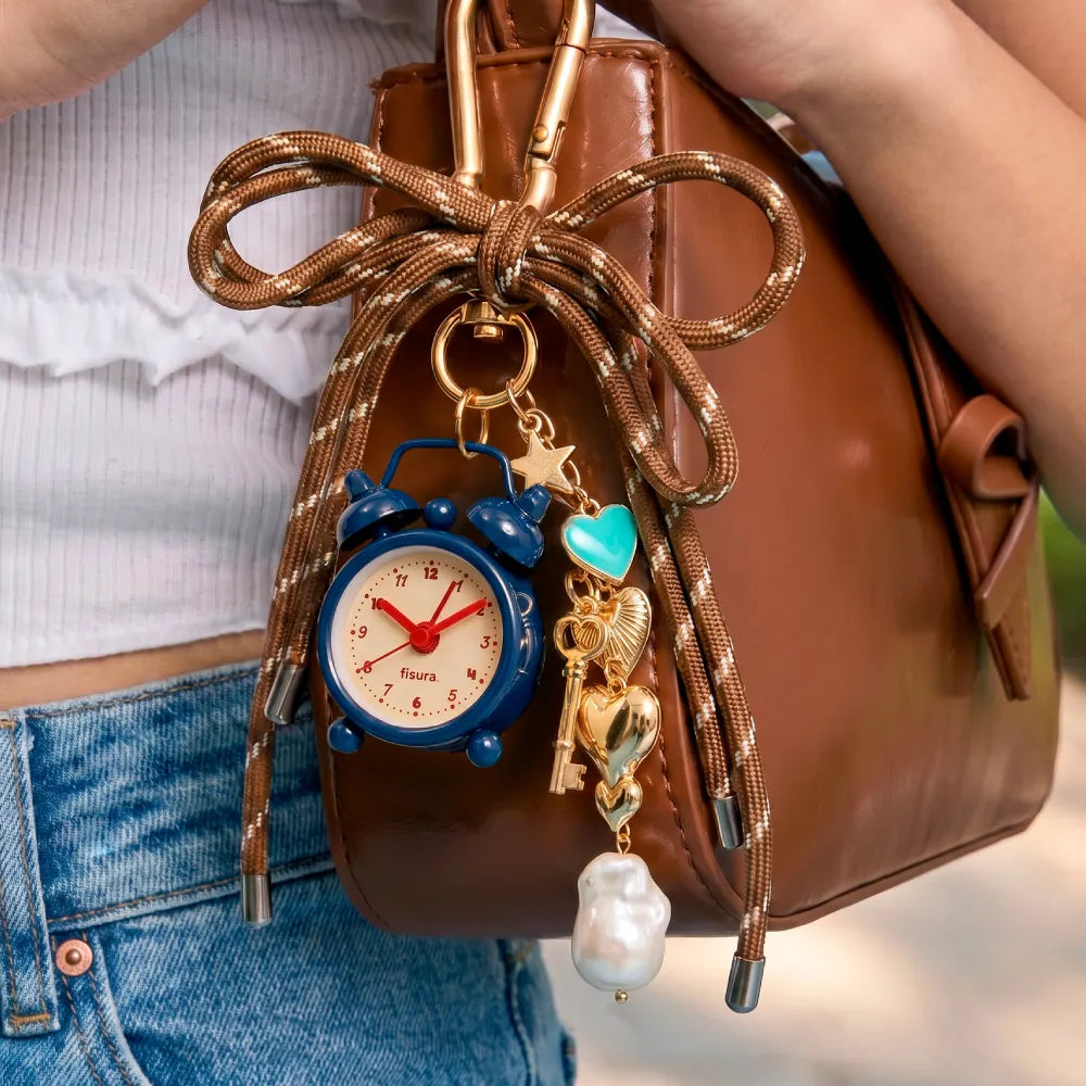 Bolso de cuero con varios complementos colgados de un mosquetón: llavero con charms dorados y un mini reloj despertador azul con las manecillas rojas 