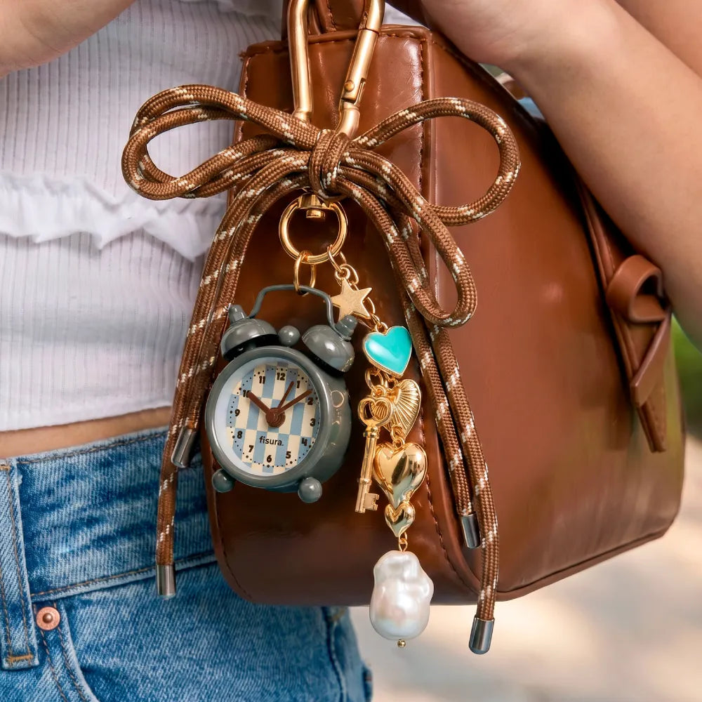 Bolso de cuero marrón con varios adornos colgados de un mosquetón, entre los que destaca un mini reloj despertador
