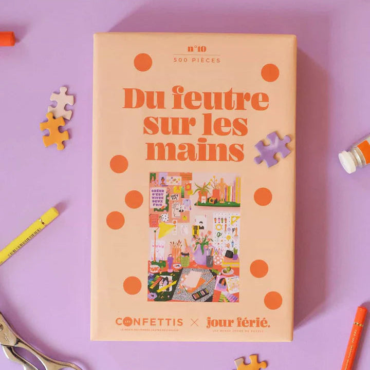 Puzzle de 500 piezas Du feutre sur les mains de Jour Férié, caja de color melocotón con ilustración de estudio creativo, rodeada de piezas sueltas y material de arte sobre fondo lila