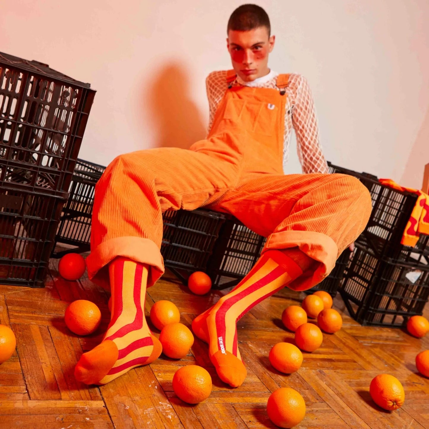 modelo posando con los calcetines de naranjas de doiy EMS sobre cajas de naranjas y con naranjas por el suelo, llevando un peto también de color naranja