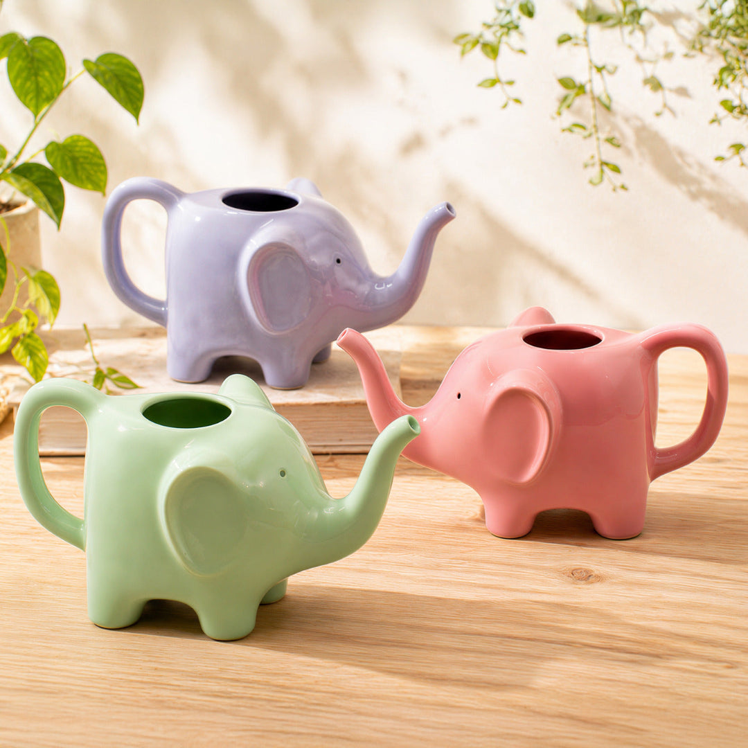 tres regaderas en forma de elefante en rosa, lila y verde encima de una superficie de madera con plantas alrededor. 