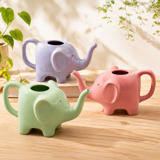 tres regaderas en forma de elefante en rosa, lila y verde encima de una superficie de madera con plantas alrededor. 