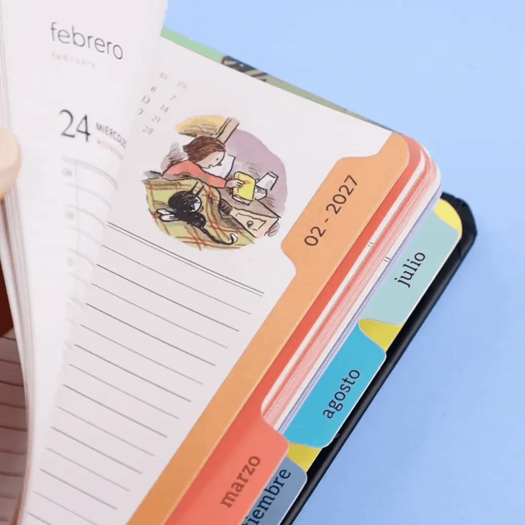 Detalle del lomo de agenda 2027 Macanudo con pestañas mensuales de colores y página de febrero con viñeta de Enriqueta