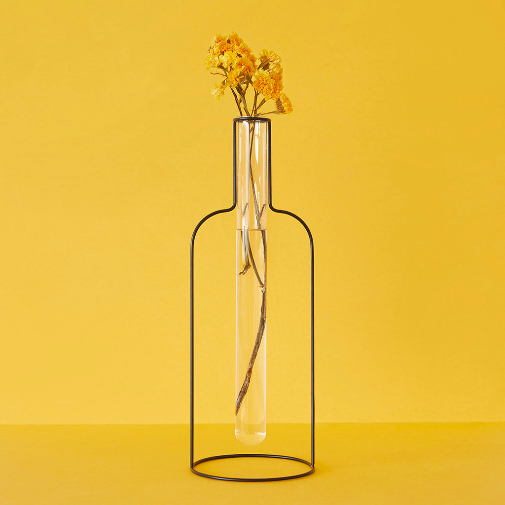 Florero con forma de silueta de botella de hierro con una probeta de cristal que contiene un tallo de flores secas con el fondo amarillo