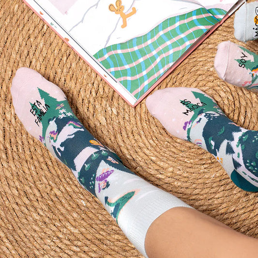 Pies sobre una alfombra de rafia y un cuento abierto cerca con los calcetines con estampado de personajes de cuento y el mensaje "esta familia es mágica"