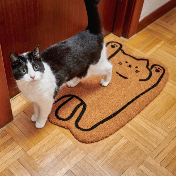 Gato blanco y negro de pie sobre un felpudo marrón con forma de gato boca arriba