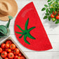 Felpudo con forma de medio tomate junto a una caja llena de tomates rojos, una tomatera, una regadera y un gorro de paja