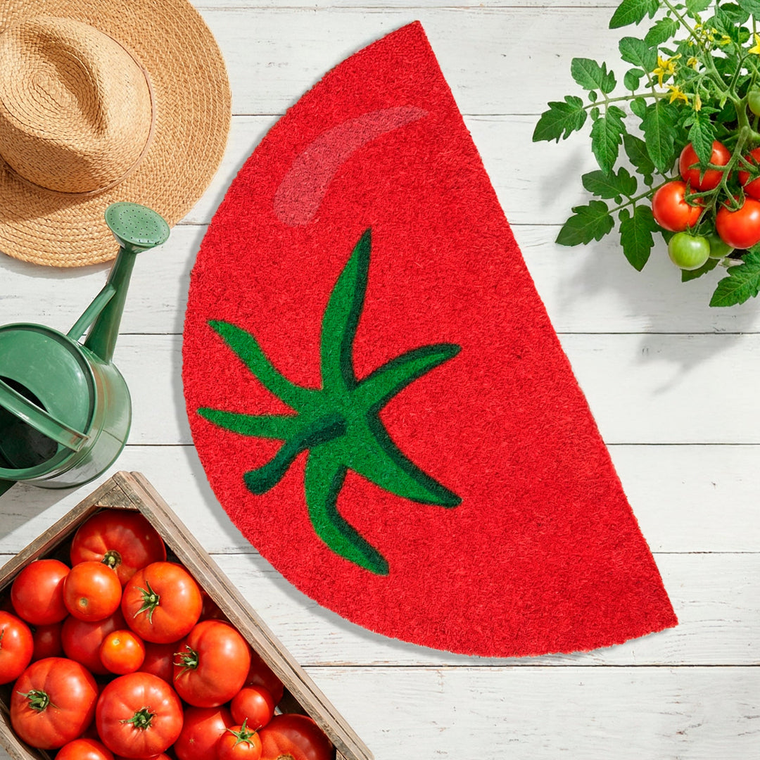 Felpudo con forma de medio tomate junto a una caja llena de tomates rojos, una tomatera, una regadera y un gorro de paja