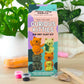 Kit DIY Gatos Curious Kitties, con caja ilustrada, arcilla de secado al aire, pinturas y herramientas sobre una mesa entre plantas