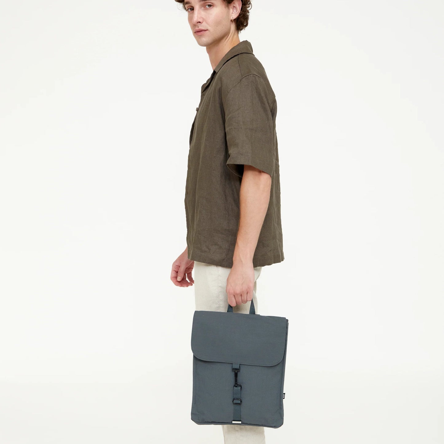 Modelo masculino de perfil sosteniendo la mochila Lefrik gris pizarra por el asa, con camisa verde y pantalón beige