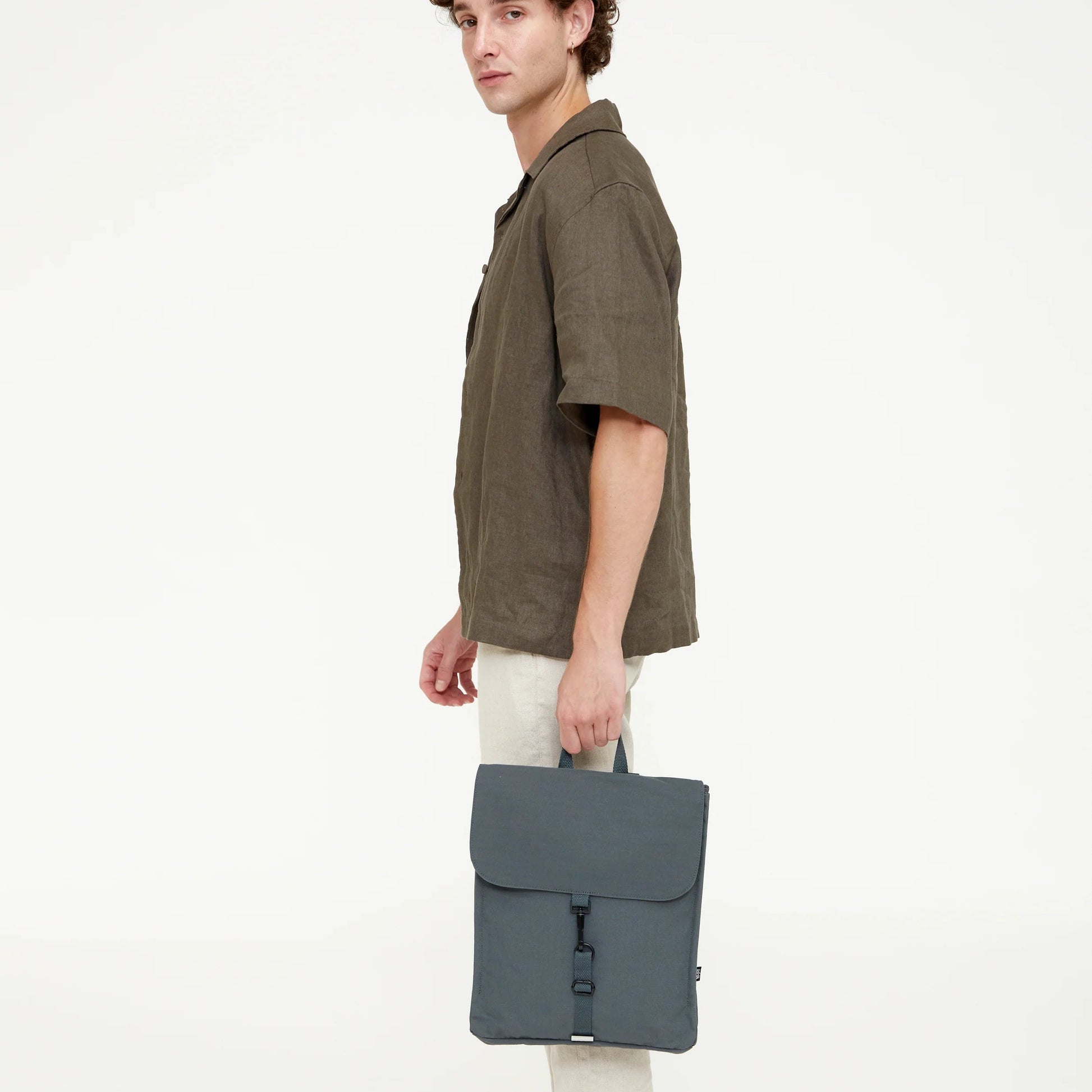 Modelo masculino de perfil sosteniendo la mochila Lefrik gris pizarra por el asa, con camisa verde y pantalón beige