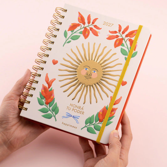 Manos sosteniendo la agenda semanal 2027 Honrá tu poder de Happimess, con tapa dura blanca decorada con un sol dorado con cara, flores rojas y hojas verdes, encuadernación de anillas doradas y goma elástica amarilla