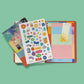 Vista cenital de los extras incluidos en la agenda Happimess 2027: láminas ilustradas, hoja de pegatinas con iconos y frases motivacionales, regla marcapáginas con sol y tapa trasera con cuadrícula de colores pastel