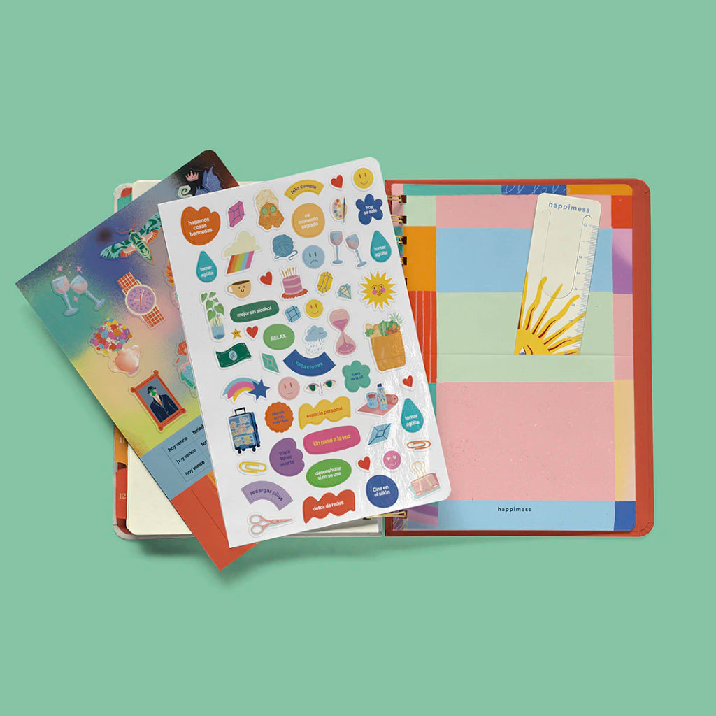 Vista cenital de los extras incluidos en la agenda Happimess 2027: láminas ilustradas, hoja de pegatinas con iconos y frases motivacionales, regla marcapáginas con sol y tapa trasera con cuadrícula de colores pastel