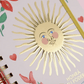 Detalle en primer plano del sol dorado con cara de la tapa de la agenda Happimess 2027 Honrá tu poder, con acabado stamping dorado y flores rojas al fondo