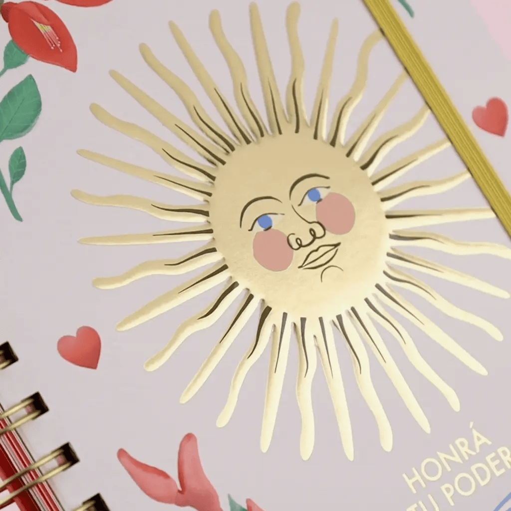 Detalle en primer plano del sol dorado con cara de la tapa de la agenda Happimess 2027 Honrá tu poder, con acabado stamping dorado y flores rojas al fondo
