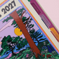 Agenda diaria 2027 Mis sueños me necesitan de Happimess con tapa dura ilustrada con un zorro en un paisaje natural de flores y árboles, encuadernación de anillas doradas y goma elástica naranja