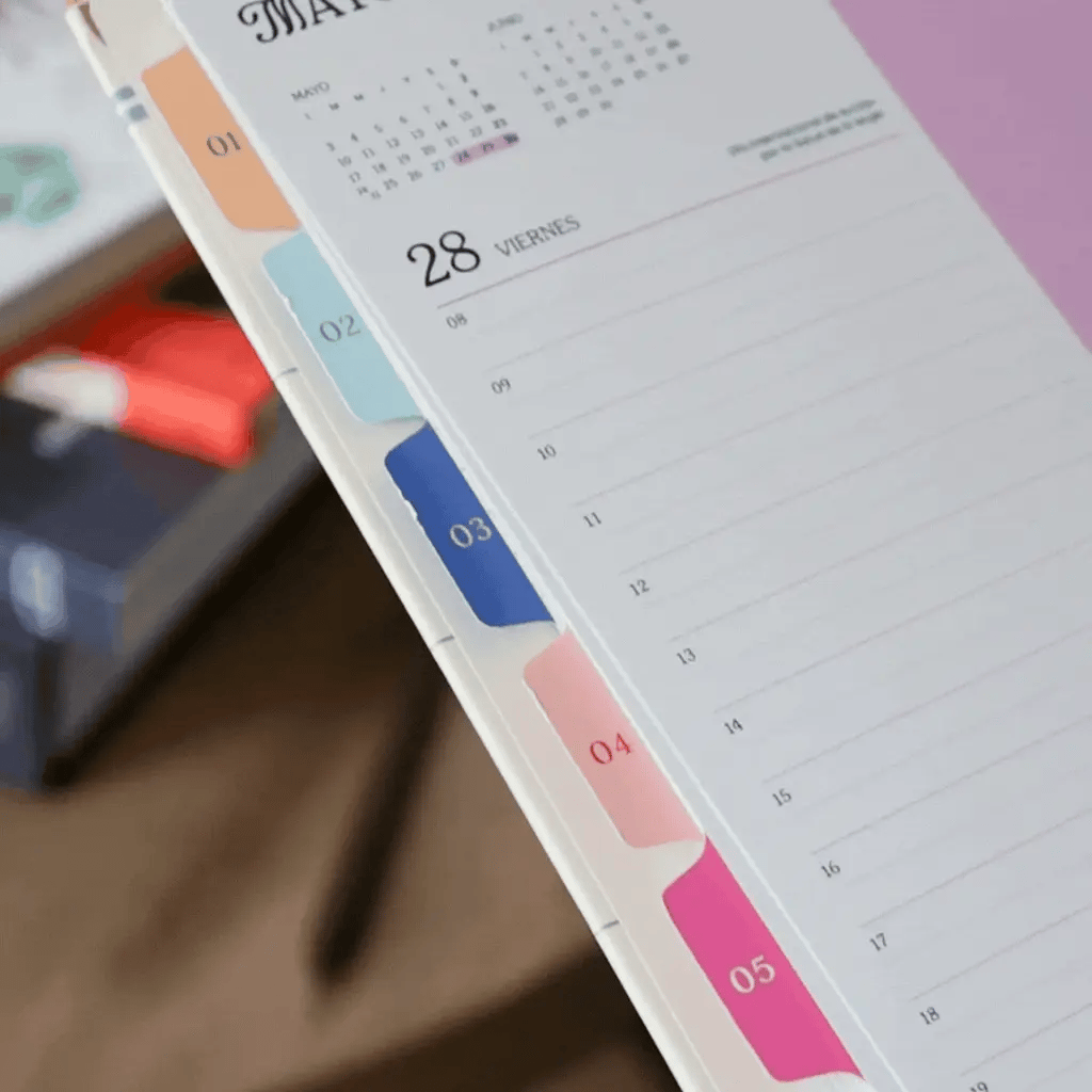 Lomo de la agenda diaria 2027 Happimess con separadores mensuales de colores numerados del 01 al 05 en tonos naranja, azul, azul marino, rosa y fucsia, con páginas diarias visibles