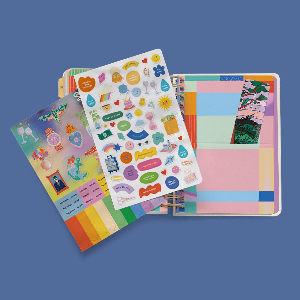 Agenda diaria 2027 Happimess abierta junto a dos hojas de pegatinas incluidas, con ilustraciones coloridas, emojis y frases motivacionales, y separadores mensuales de colores pastel visibles al fondo