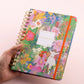 Agenda semanal 2027 Happimess Primavera con tapa dura floral en tonos verdes rosas y lilas con espiral dorada y goma naranja sostenida en mano fondo rosa