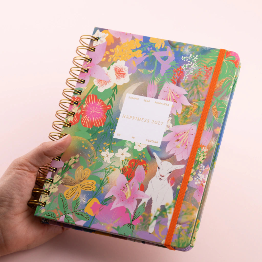 Agenda semanal 2027 Happimess Primavera con tapa dura floral en tonos verdes rosas y lilas con espiral dorada y goma naranja sostenida en mano fondo rosa