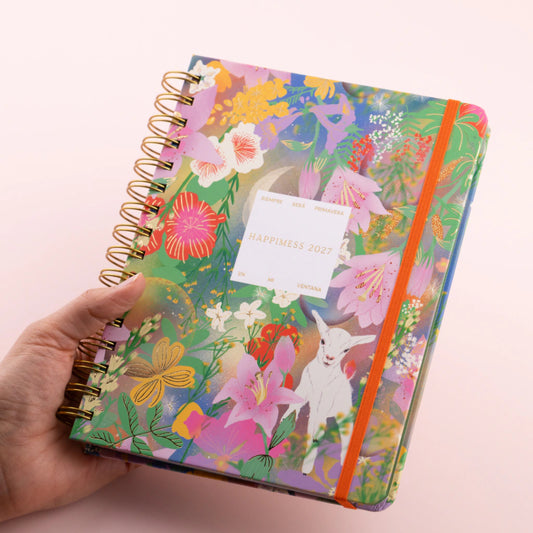Agenda semanal 2027 Happimess Primavera con tapa dura floral en tonos verdes rosas y lilas con espiral dorada y goma naranja sostenida en mano fondo rosa
