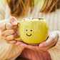 Taza amarilla con carita sonriente aguantada por unas manos 